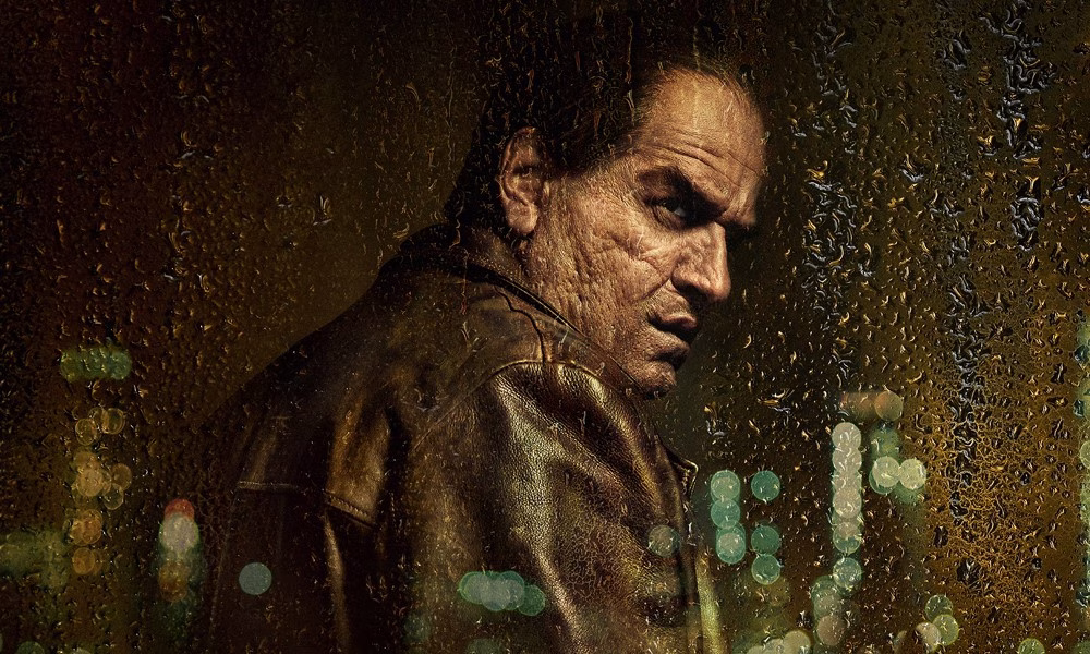 A man with a scarred face looks over his shoulder at a rain-spattered window, through which we can see the reflection of a city skyline.