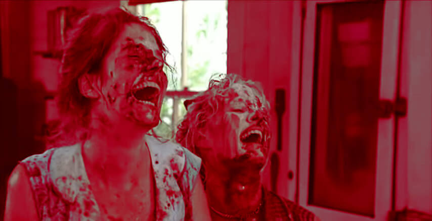 Two women, covered in chocolate frosting and flour, laugh heartily. The image has a red cast to make the chocolate look like blood.