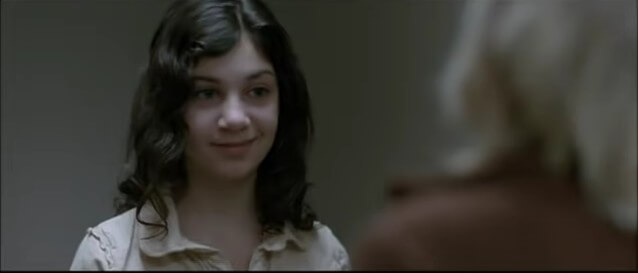 In a doorway, a dark-haired young girl smirks as she talks to a boy with shaggy blond hair.