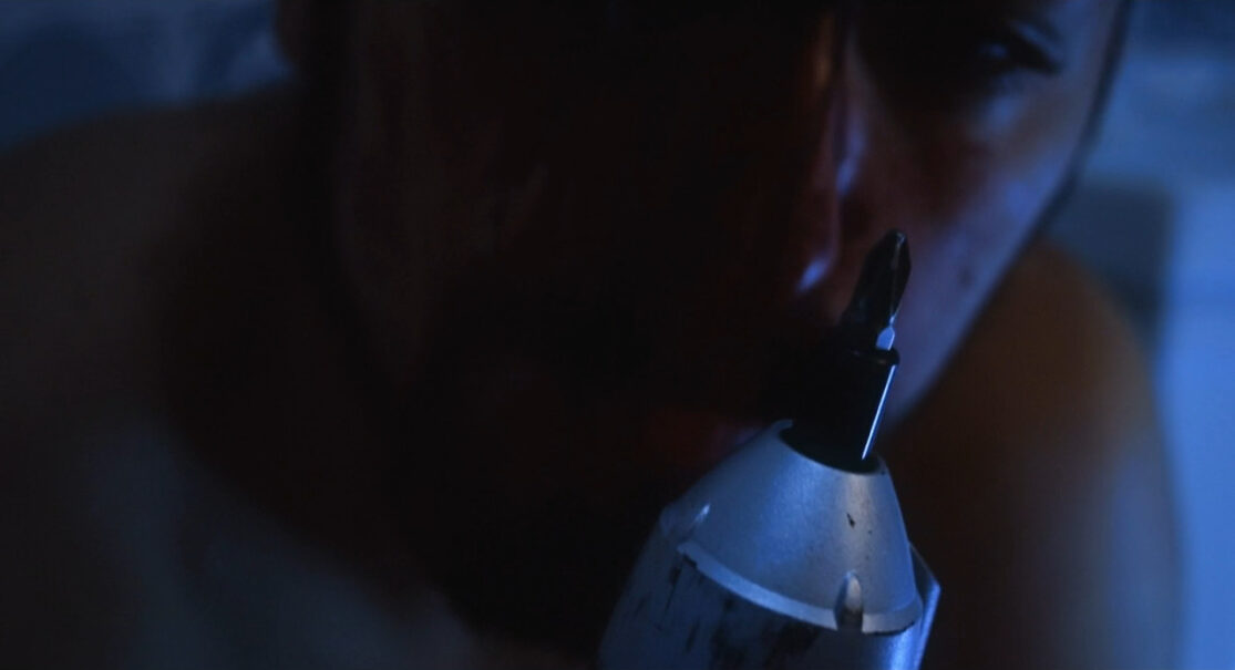 A man, shown close up in low light, contemplates using a drill with a Phillips-head screwdriver bit.