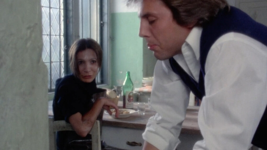 A woman, eats at a table while a man prepares to vomit in the foreground.