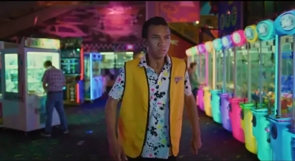 In a scene from I Saw the TV Glow, a teenage boylooks anxious among the colored lights of the arcade where he works.